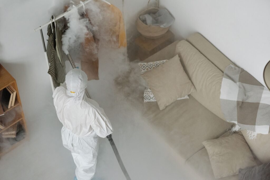 A worker in protective gear performs fumigation in a living room, using smoke to sanitize surfaces.